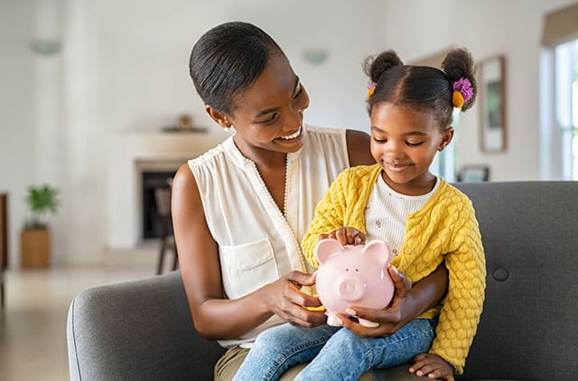 A mother looks happily at her child, while the child holds a piggy bank.