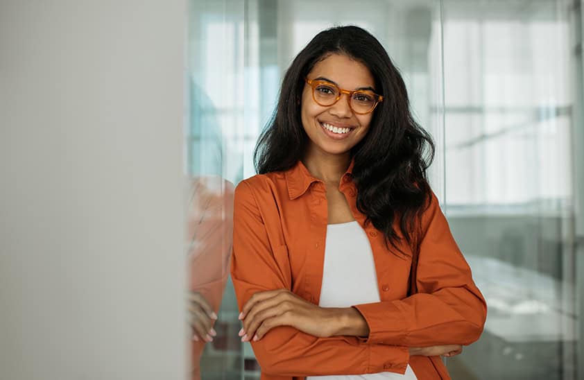 A woman wearing an orange jacket and glasses leaning against a wall.