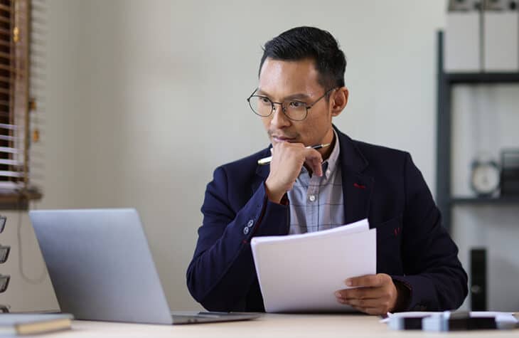 A man wearing glasses looking seriously at a laptop screen and holding documents.