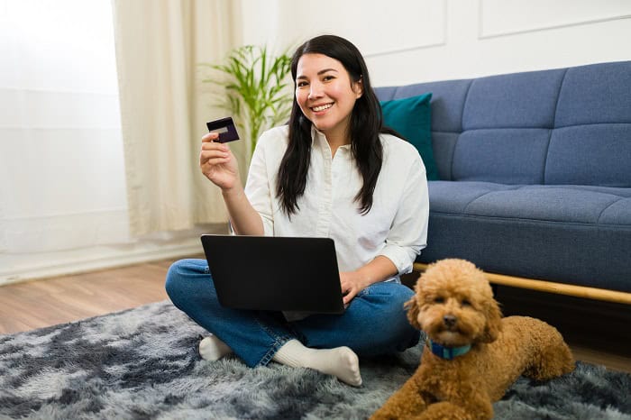 Woman sitting on the floor with laptop and credit card, pet dog beside her, in a bright, comfortable living room setting