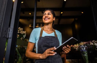 A small business owner holding a tablet while looking up and ahead while smiling.