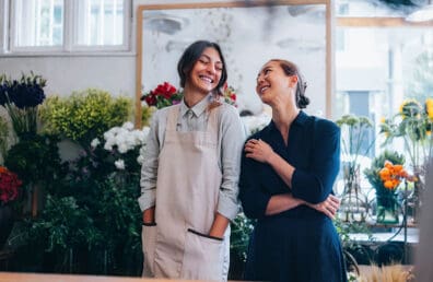 Two female florists smile while chatting with each other in a store with blooming flowers.