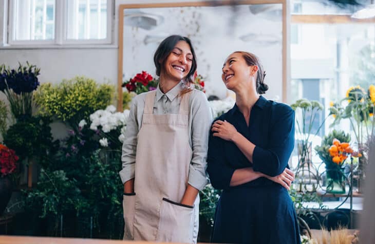 Two female florists smile while chatting with each other in a store with blooming flowers.