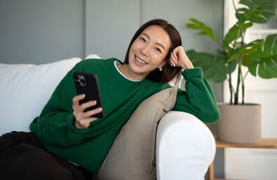 Young woman in a green sweater sits comfortably on a sofa at home, smiling at the camera while holding a smartphone, relaxed and enjoying leisure time indoors