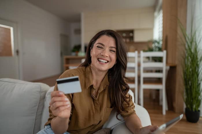 Happy mid-30s woman smiling at home, holding a credit card and tablet while enjoying convenient online shopping, secure e-commerce and modern digital banking